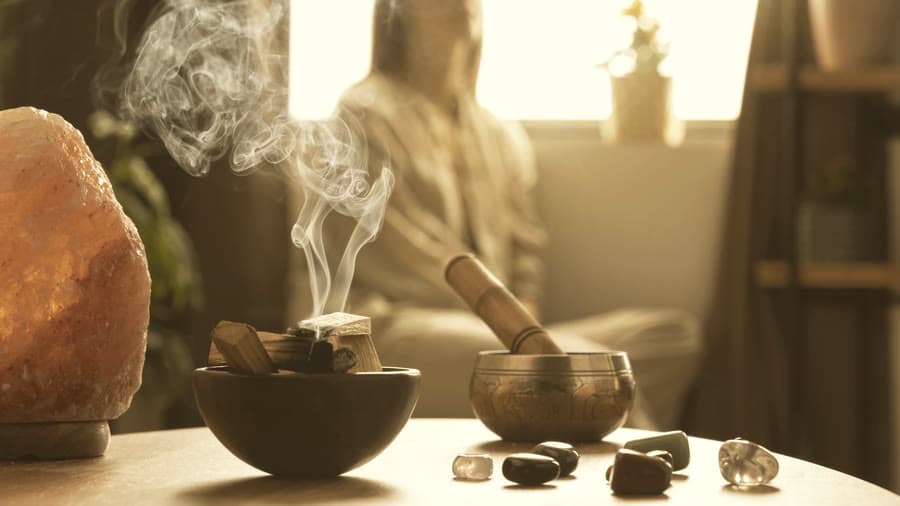 Palo santo humeando en un cuenco de madera, junto a un cuenco tibetano, cuarzos y una lámpara de sal, sobre una mesa iluminada por la ventana