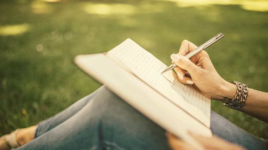 Una mujer sentada sobre el césped escribe en un cuaderno abierto sobre sus rodillas