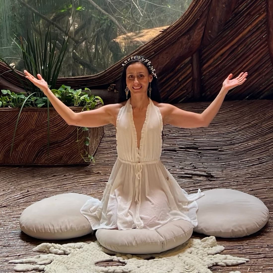 Nata Oro Gon sonriendo con los brazos abiertos, vestida de blanco y sentada sobre cojines de meditación, en una cabaña de madera